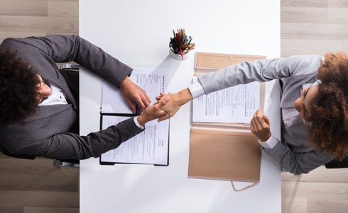 Two individuals shaking hands over a table that has papers on it
