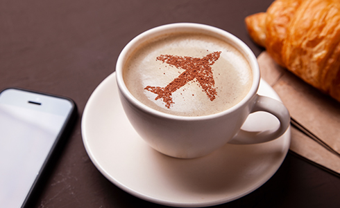 Image of a coffee cup in an airport cafe, with an airplane design on top next to a cell phone and a crossaint